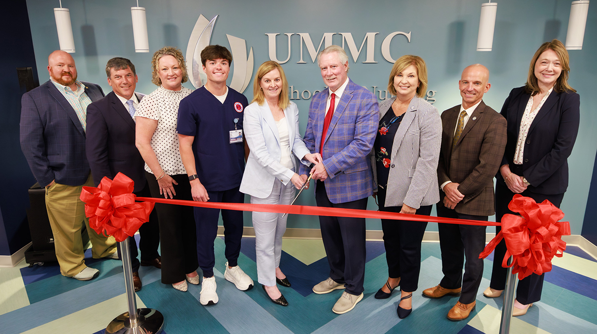 A woman and a man use a giant pair of scissors to cut a red ribbon in front of a wall sign reading 'UMMC School of Nursing' as a group of people look on.