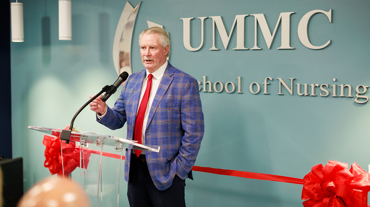 A man in a blue plaid suit speaks into a microphone in front of a sign reading 'UMMC School of Nursing.'
