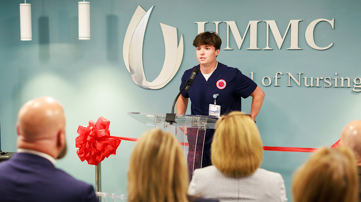 A young man wearing medical scrubs speaks to a crowd during a ceremony.