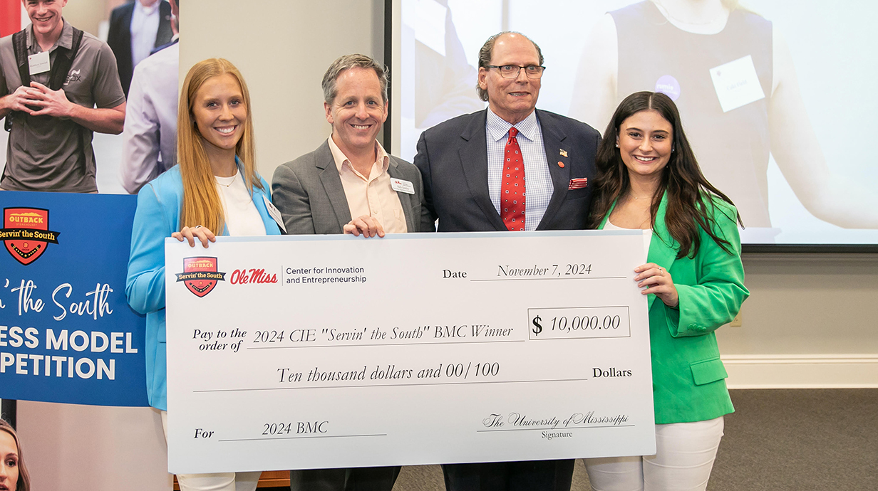 Two young women and two men hold a giant check at the front of a conference hall.