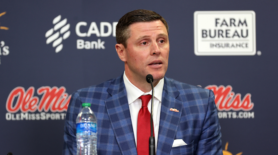A man wearing a blue plaid suit and a red tie speaks into a microphone in front of a printed backdrop.