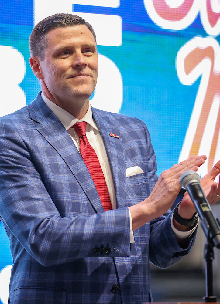 A man wearing a blue plaid suit applauds in front of a projection screen.