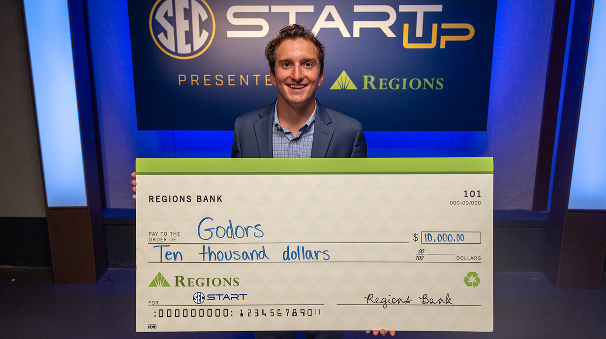 A young man wearing a suit holds a giant check for $10,000.