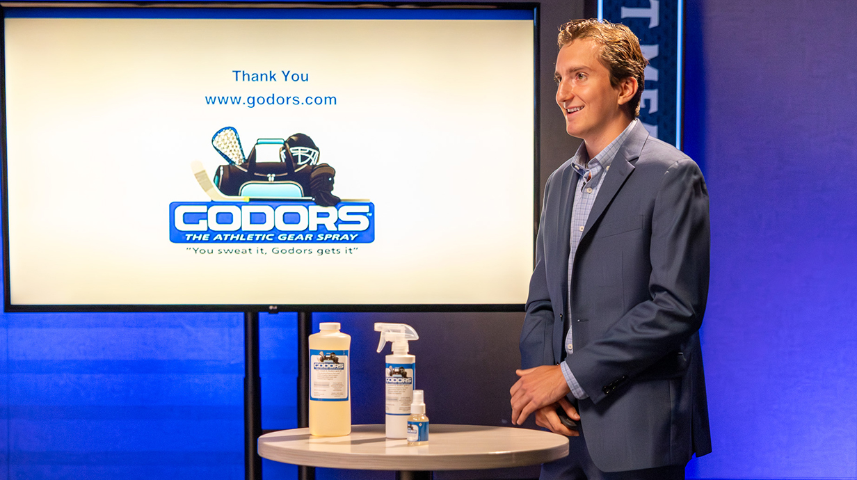 A young man wearing a suit displays cleaning products on a table in front of a television bearing an ad for Godors.