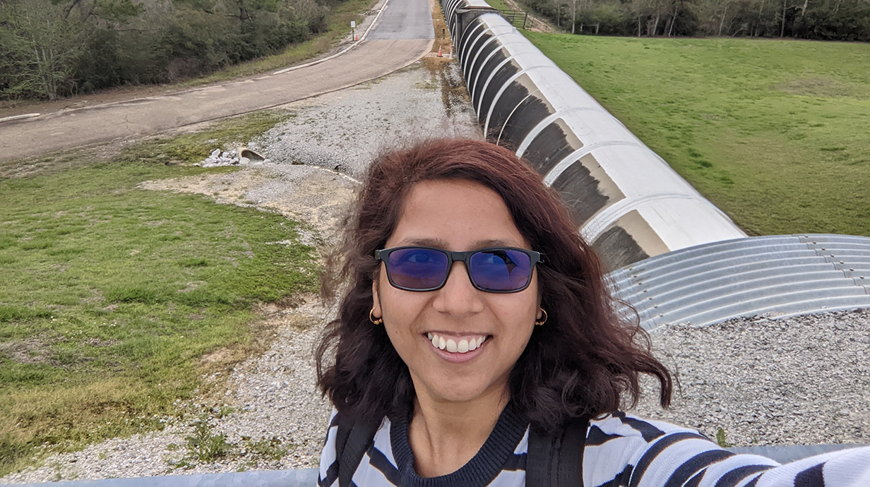 A woman wearing sunglasses takes a selfie standing in front of a giant tube half-buried in the earth of a grassy field.