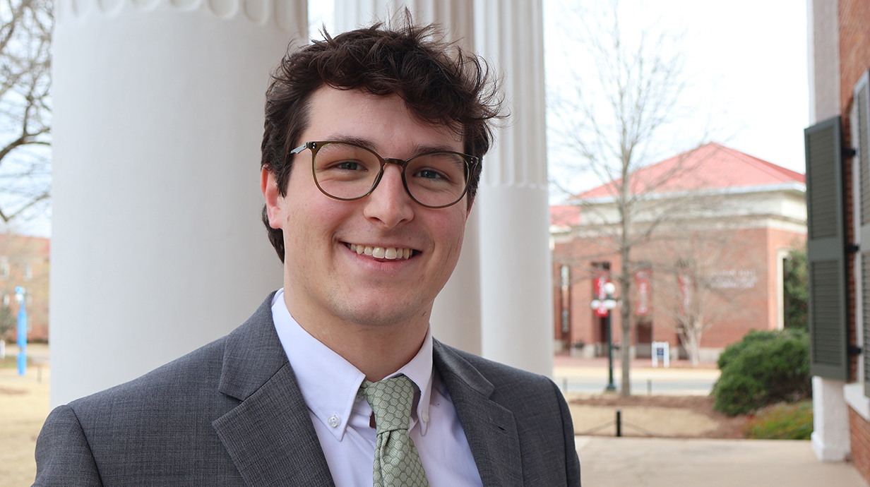 A young man wearing a gray suit stands next to several large, white columns outside a building.