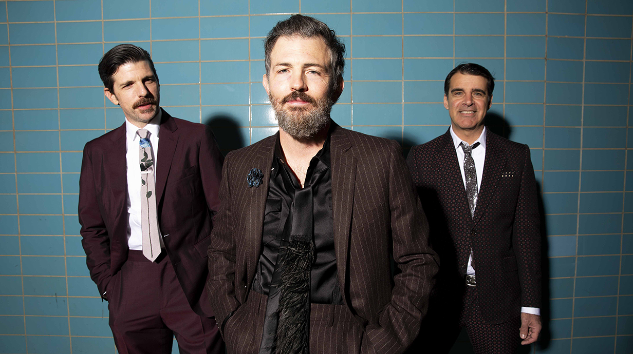 Three men wearing dark suits stand in front of a wall lined with aqua-colored ceramic tiles.