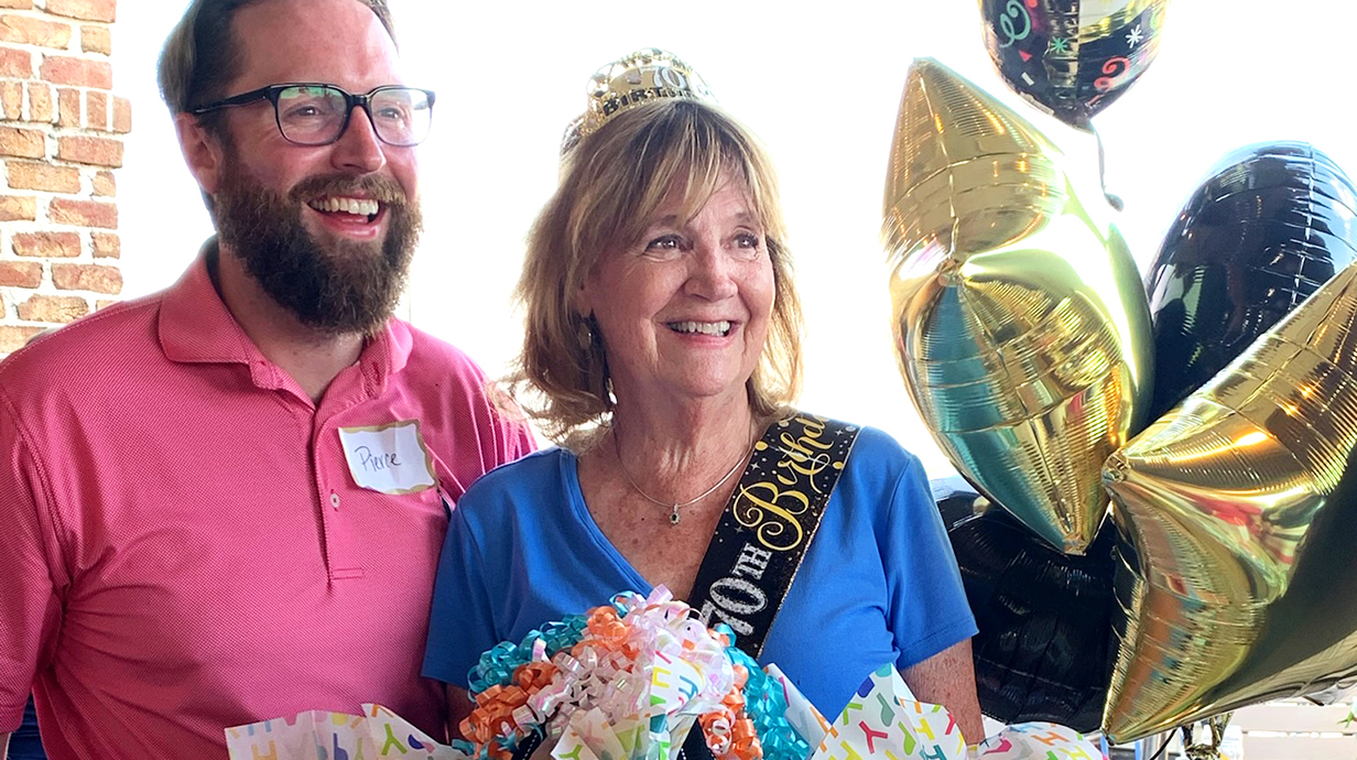 A man and a woman wearing a tiara stand among balloons and wrapping paper at a birthday party.