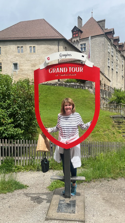 A woman standing in front of a large stone building looks through an opening in a sign reading 'Grand Tour of Switzerland.'