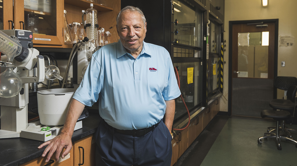 A man wearing a light blue polo shirt with stitching reading 'Ole Miss Pharmacy' stands in a laboratory.