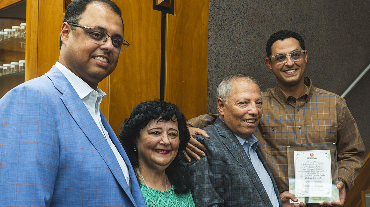 Three men, one holding a framed certificate, and a woman, stand together on a stairway.