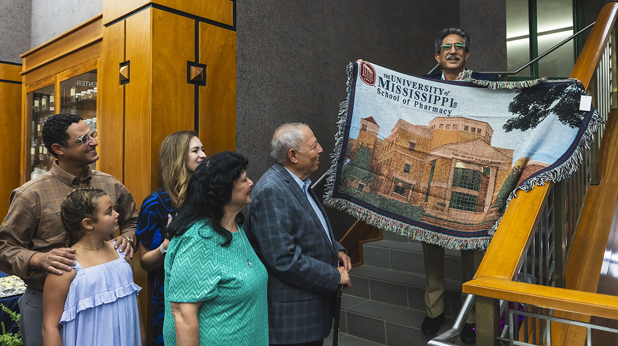 A man standing on a staircase displays a printed throw as a group of people look on.