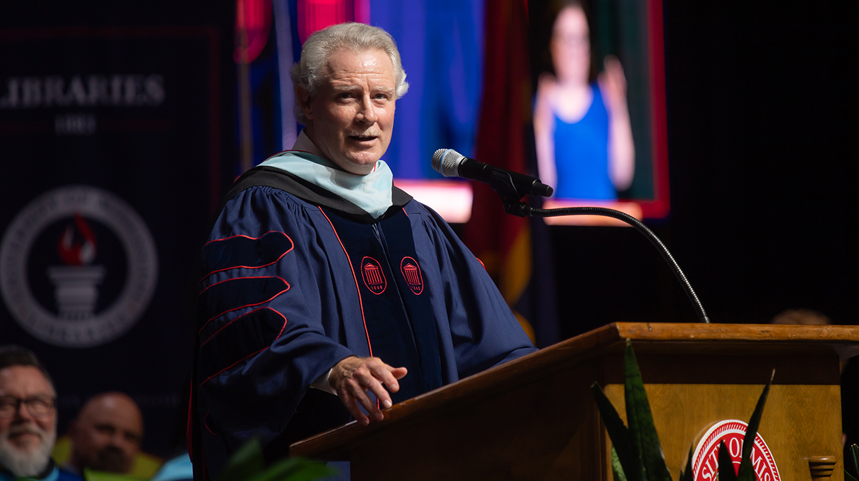 A man wearing academic regalia speaks to a crowd from a podium.