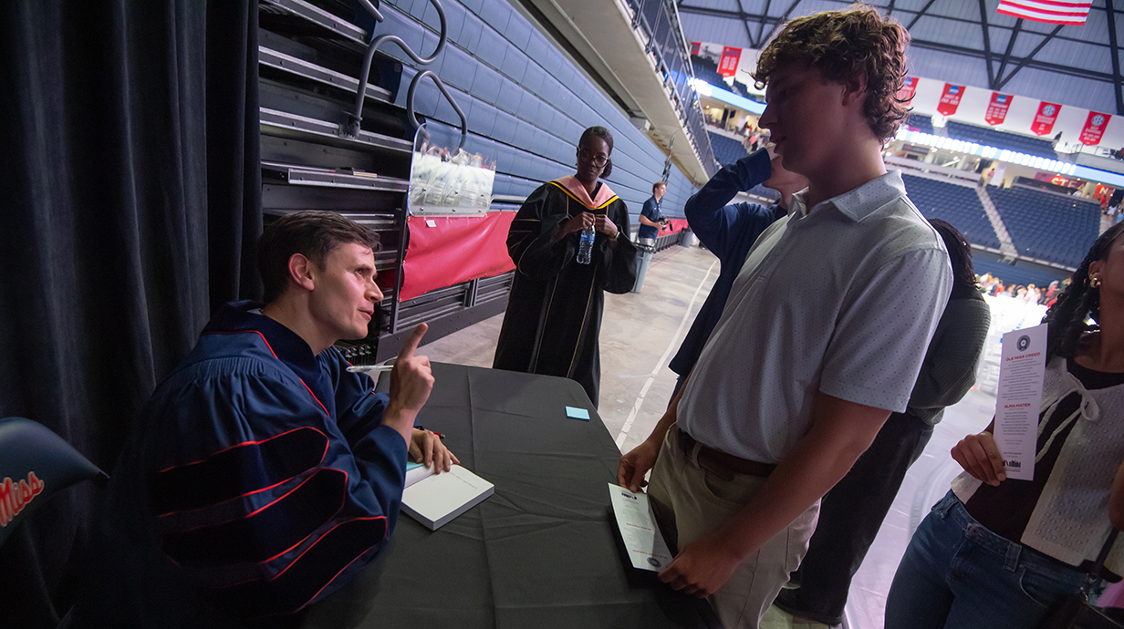 A man sitting at a table chats with a young man as he signs a book for him.