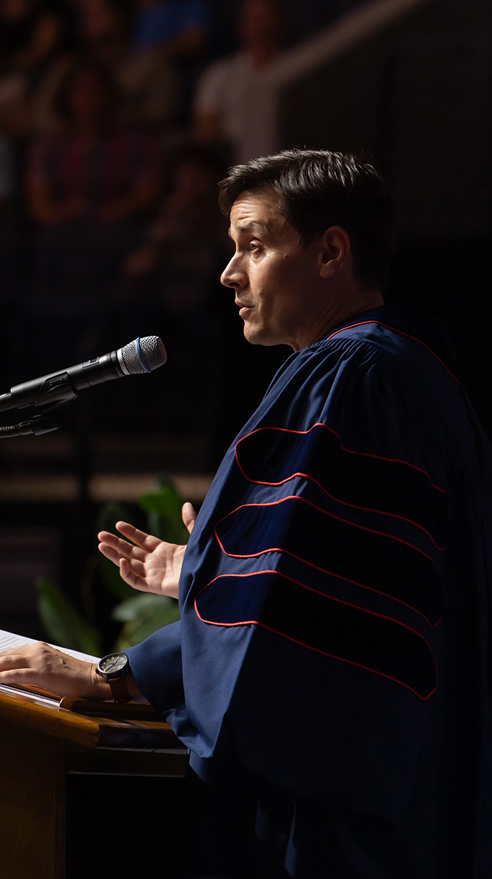 A man wearing academic regalia speaks from a podium.