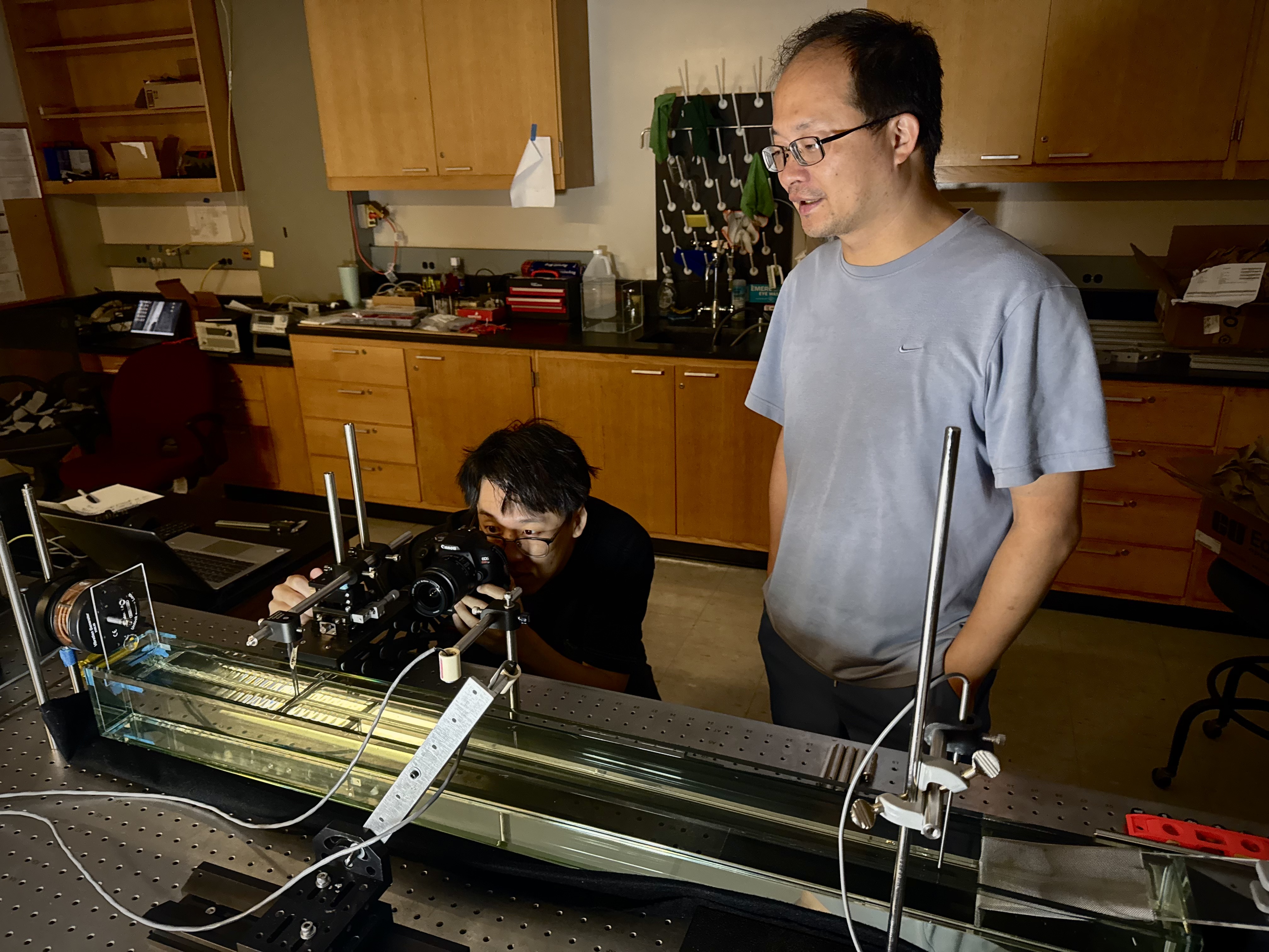 Likun Zhang watches as Zhengwu Wang looks through a camera at their experiment, where water is passing through a barrier at various heights in a controlled tank. 
