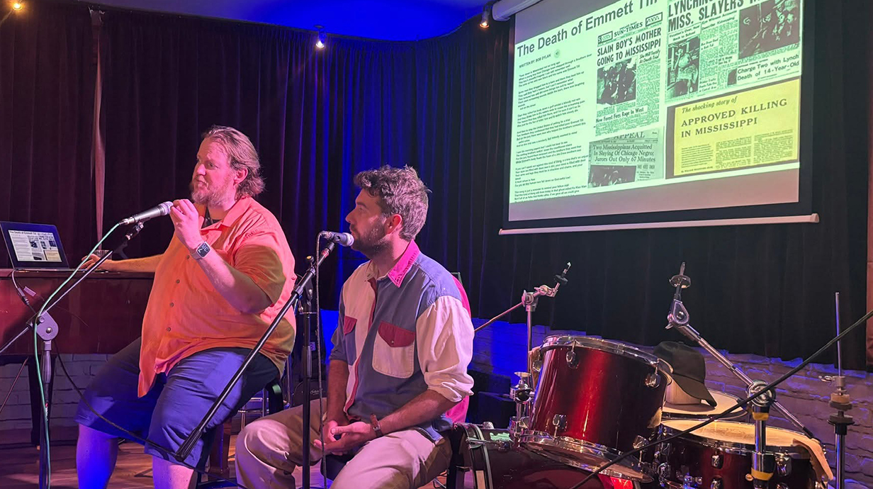 Two men sit in front of a drum kit on a stage with a projection screen behind.