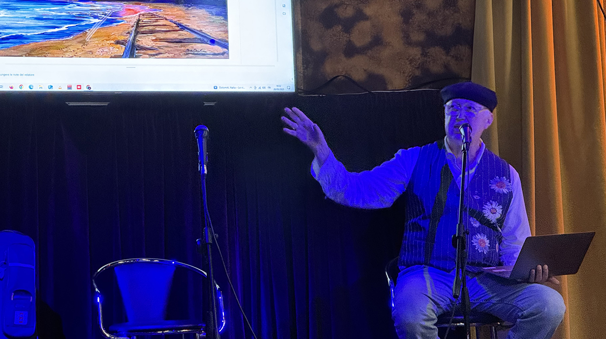 A man sitting in a darkened room holds a laptop and gestures with his other hand to a projection screen behind him.
