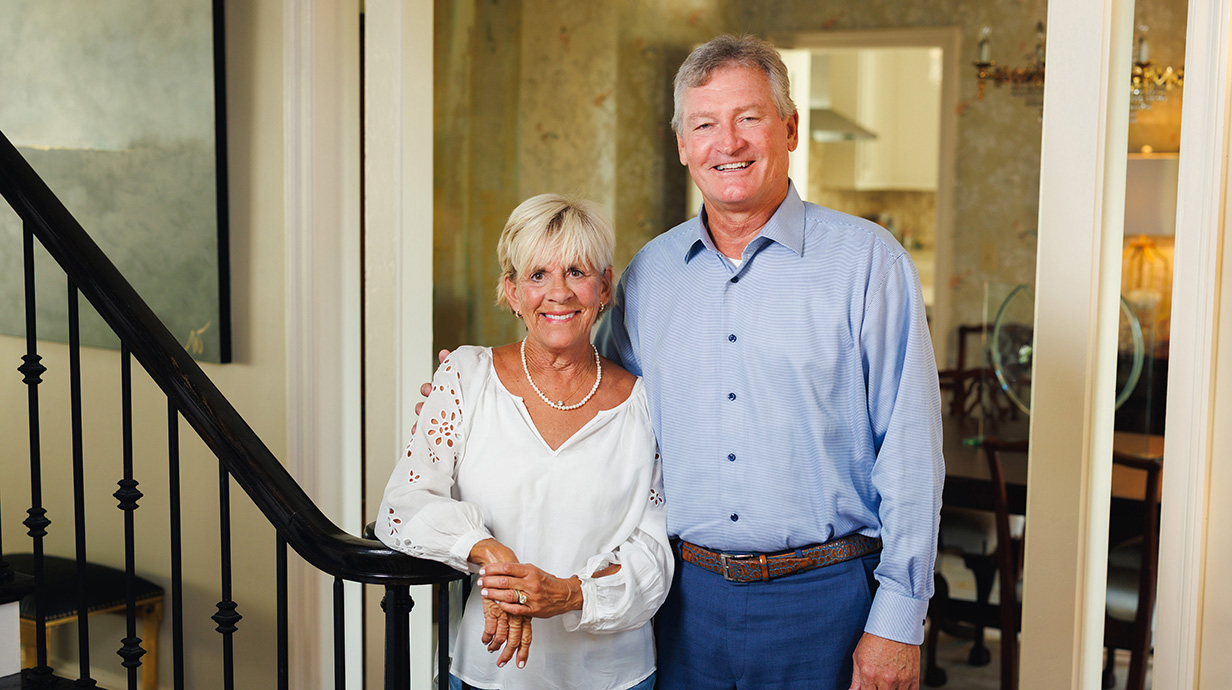 A woman and a man stand in an entry hallway, with the woman leaning against a stair railing.