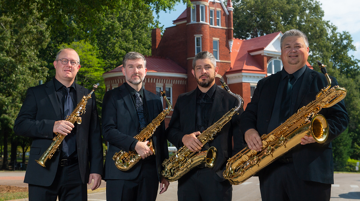 Four men, all dressed in black and holding saxophones, stand in front of a Gothic red brick building.