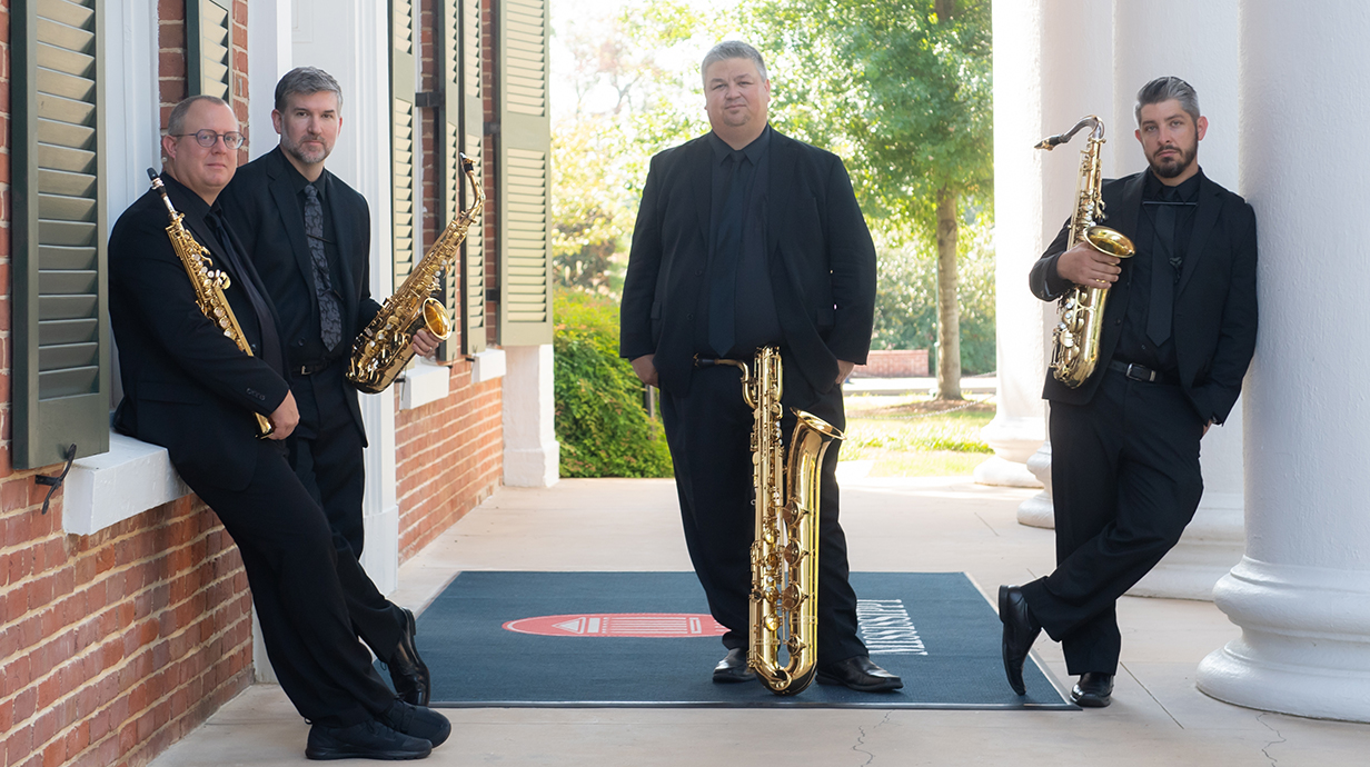 Four men, all dressed in black, hold saxophones while standing on a covered porch.