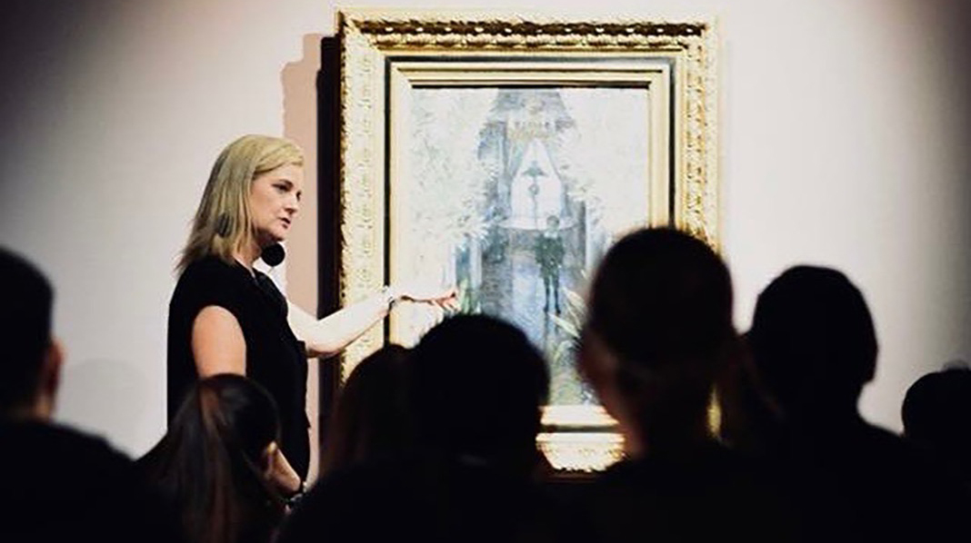 A woman points to a painting in front of a group of people in a gallery.