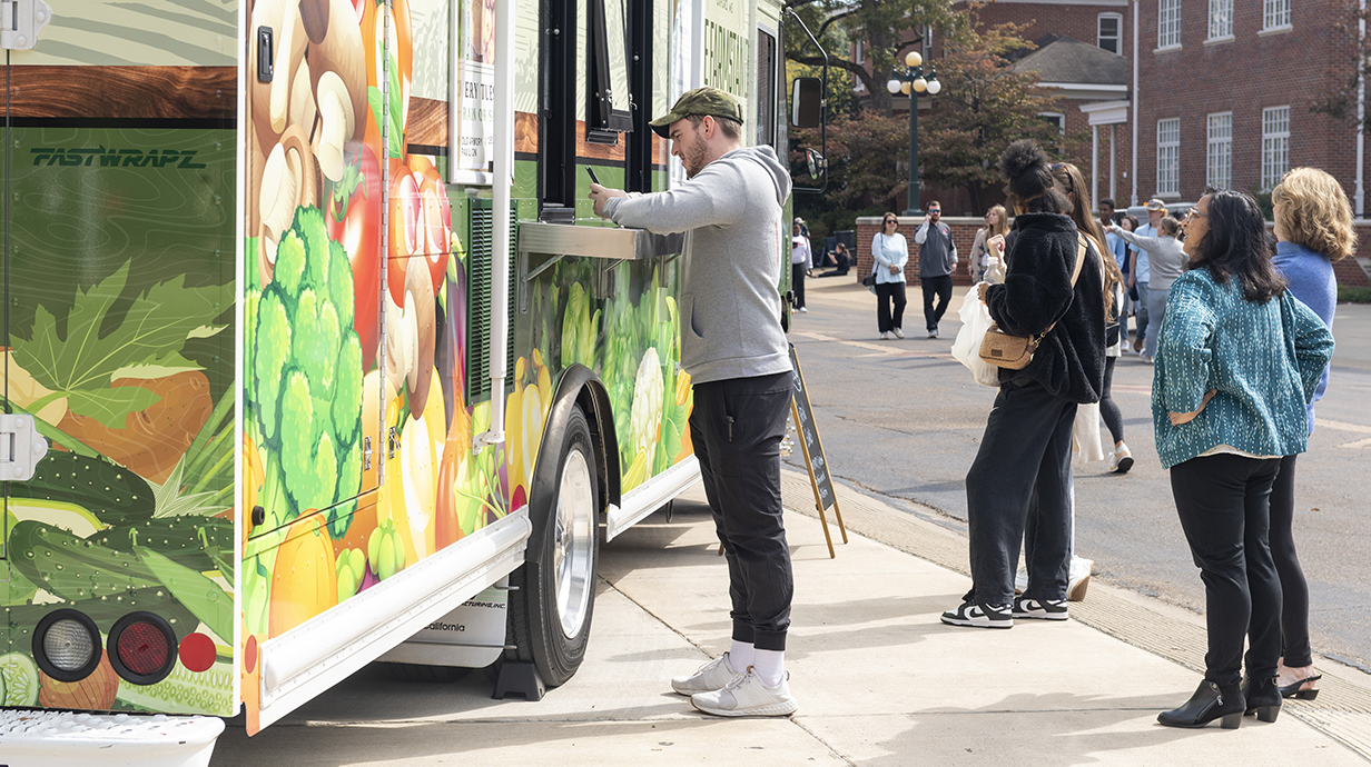Several young people line up outside a food truck while a young man leans against the counter to order.