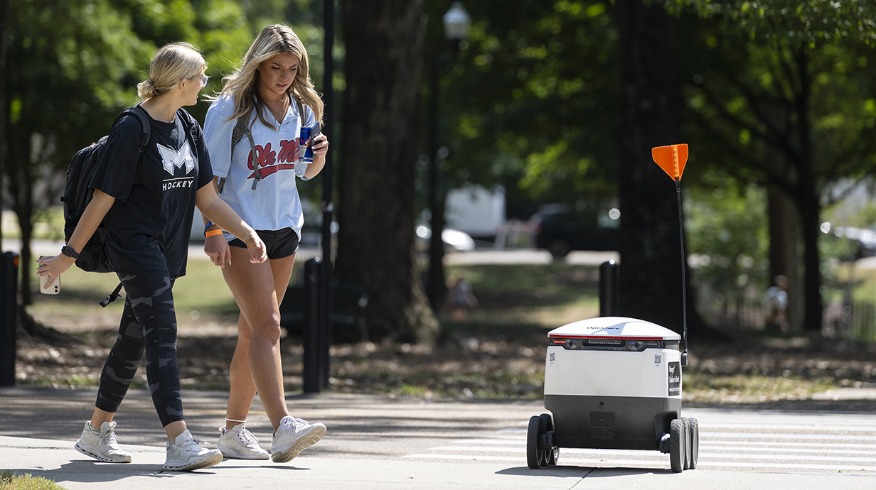 Two young women walk past a food delivery robot on a sidewalk in a tree-lined park.