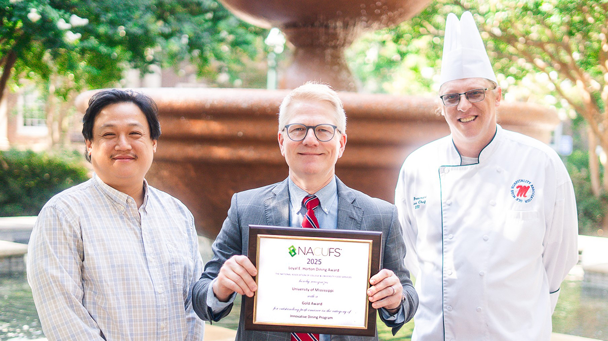 Three men – one wearing a chef's uniform and hat – hold an award certificate in front of an outdoor fountain.