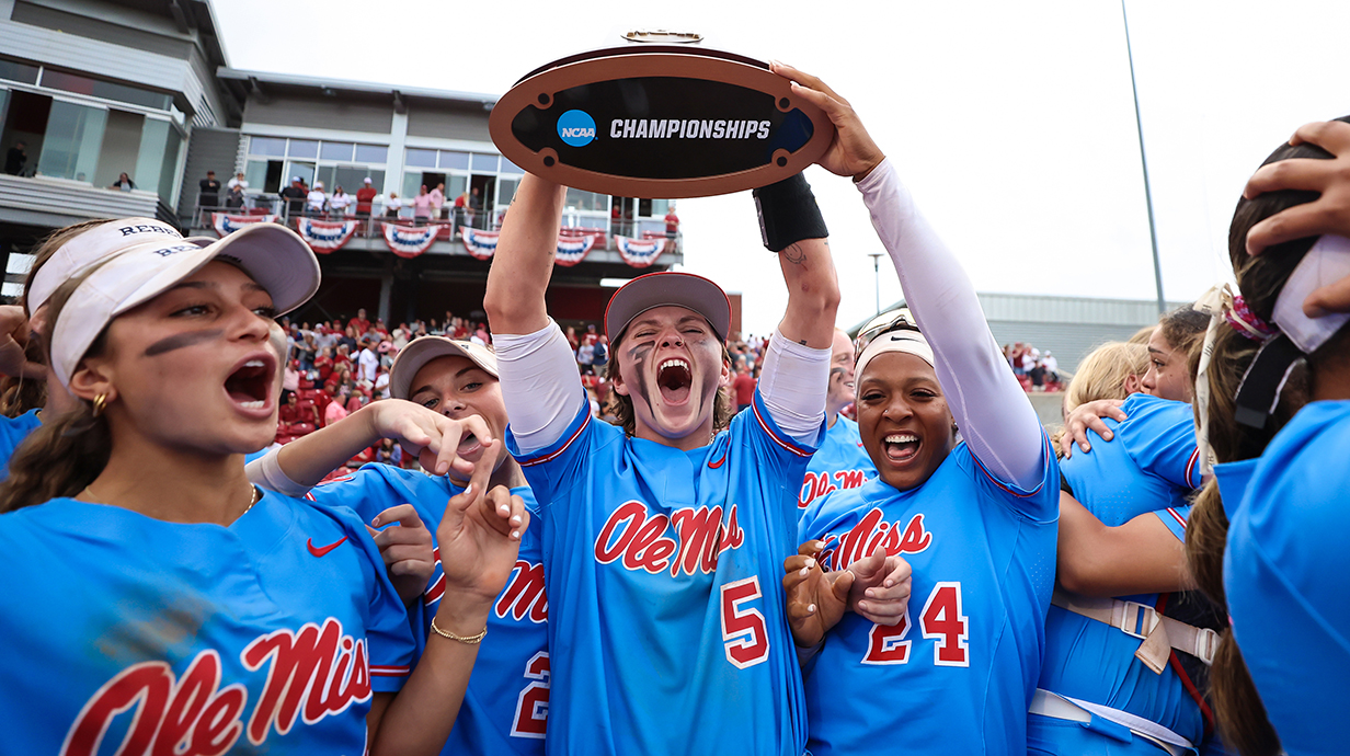 A women's softball team celebrates while raising a trophy overhead.
