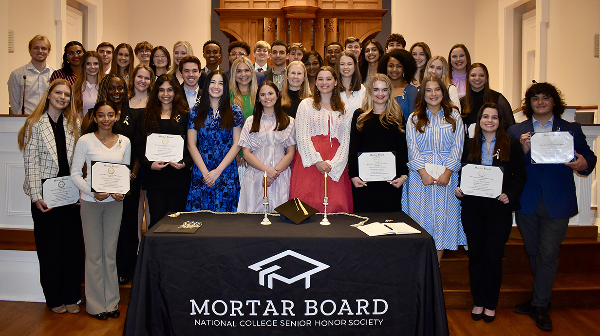 A large group of young people, all holding certificates, stand at the front of a chapel behind a table with a drape reading 'Mortar Board.'