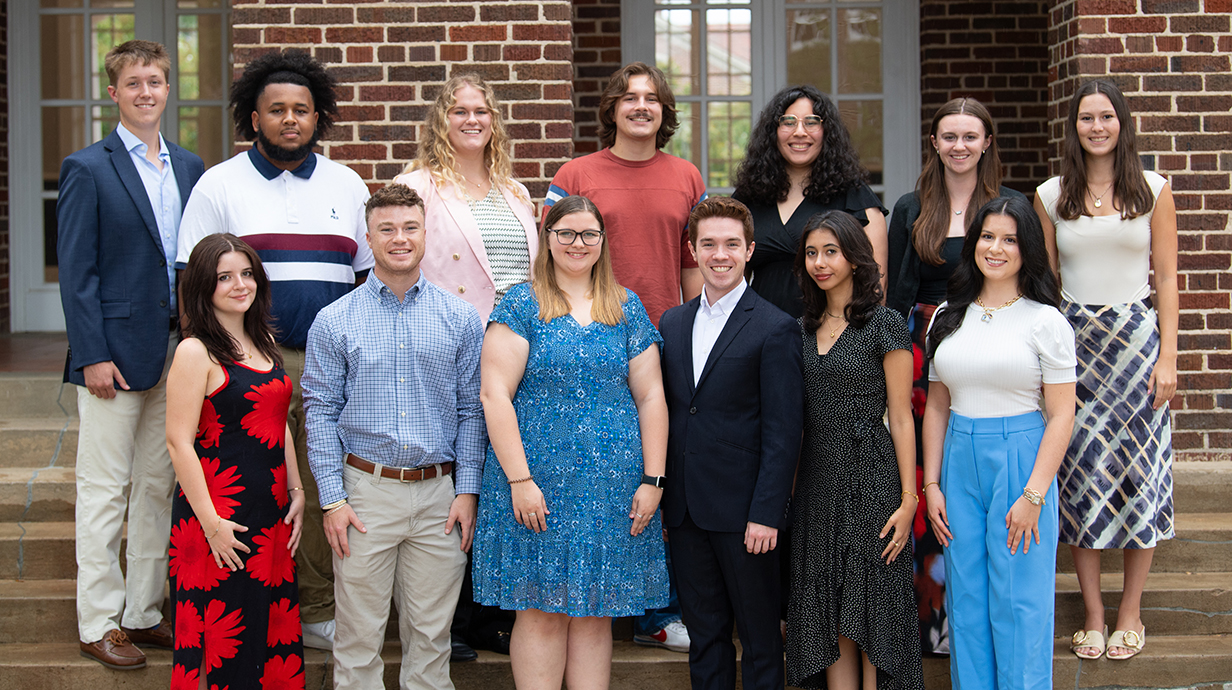 A large group of young men and women gather for a photo on concrete steps in front of a brick building.