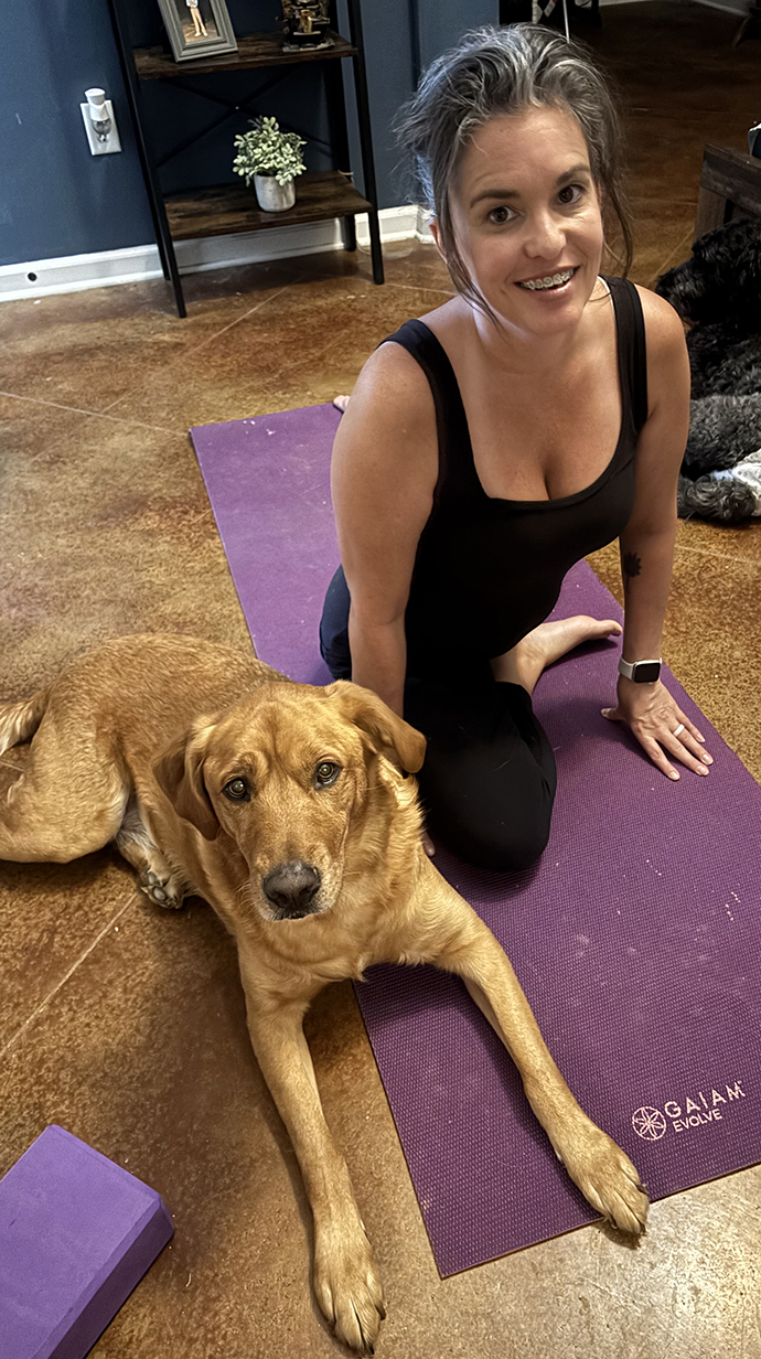 A woman sits on a yoga mat with a dog.