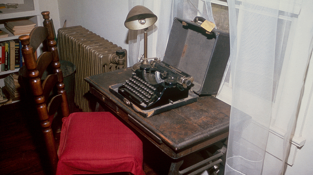 A typewriter sits on a desk in front of a chair upholstered with red fabric.