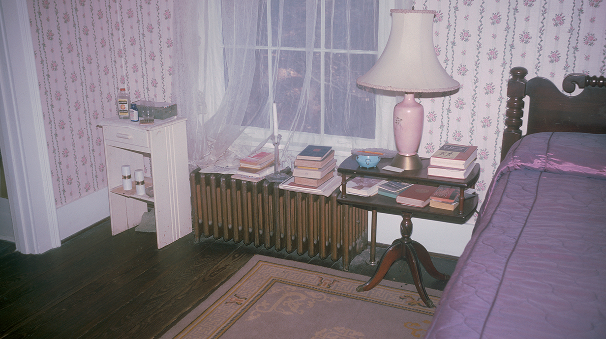 Piles of books sit on tables near a bed in a bedroom.