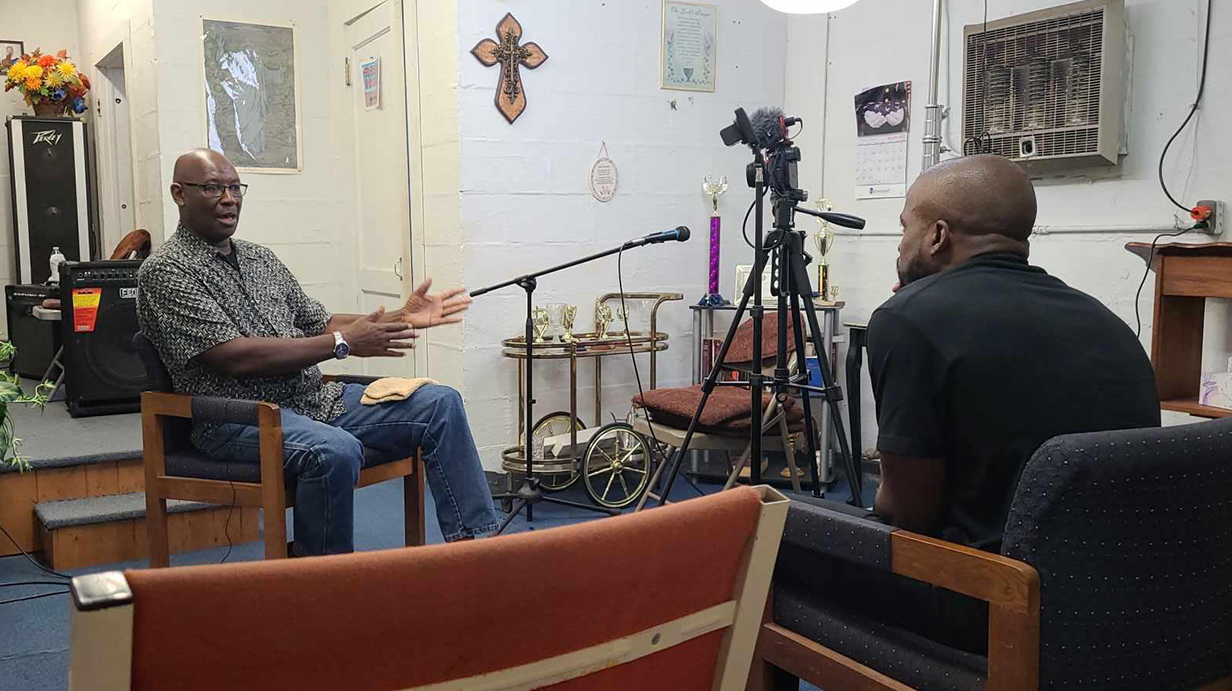 A man gestures as he talks to another man while a video camera records the scene.