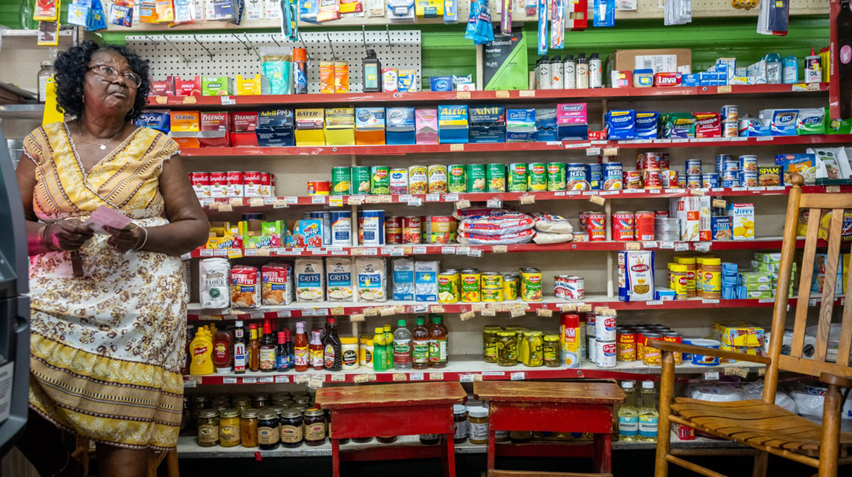 A woman stands at the counter of a small grocery store with shelves of colorful goods arranged behind her.