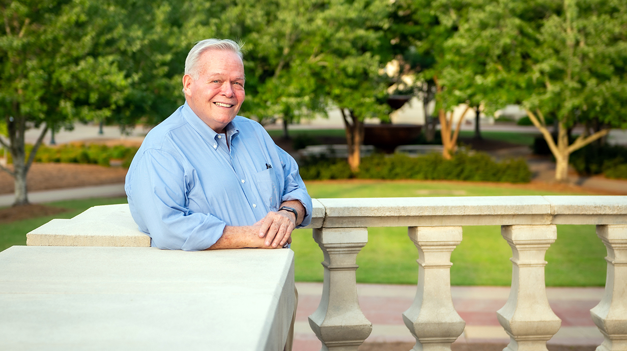 A smiling man leans against a concrete wall in a tree-lined park.