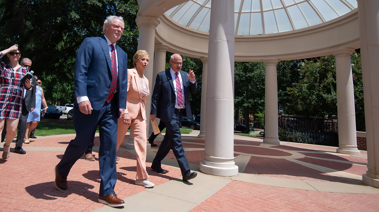 Two men and a woman walk past a columned porch as they stroll along a brick walkway.