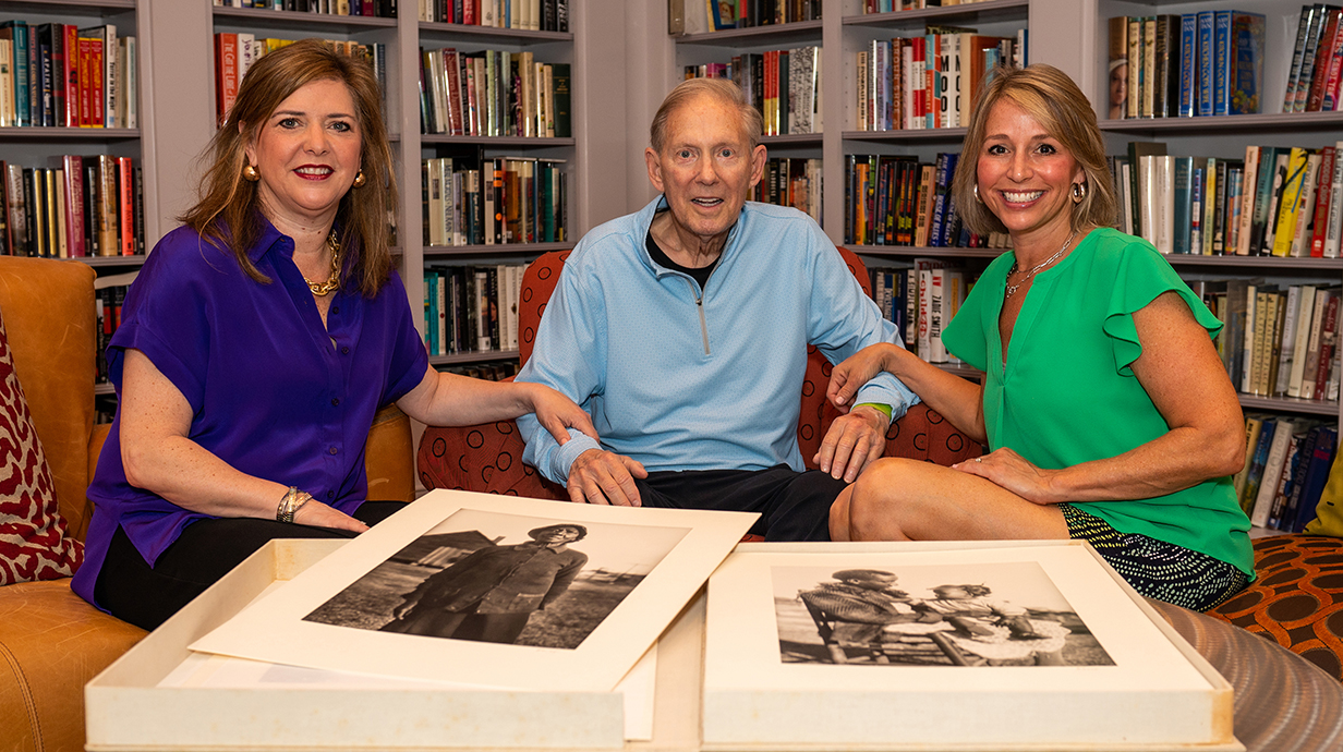 A man and two women sit on couches surrounded by bookcases and photographs.
