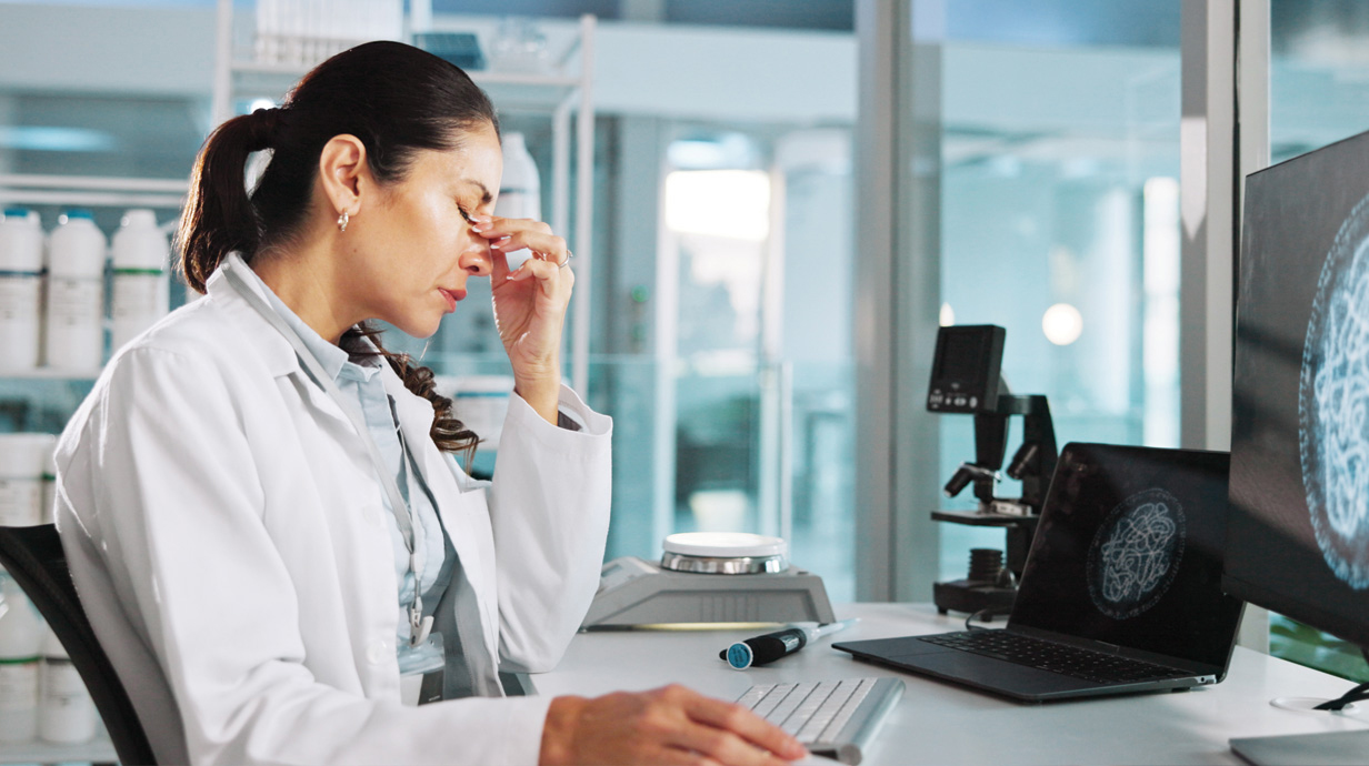 A woman sitting at a desk in a laboratory holds her face in her hand to deal with stress.