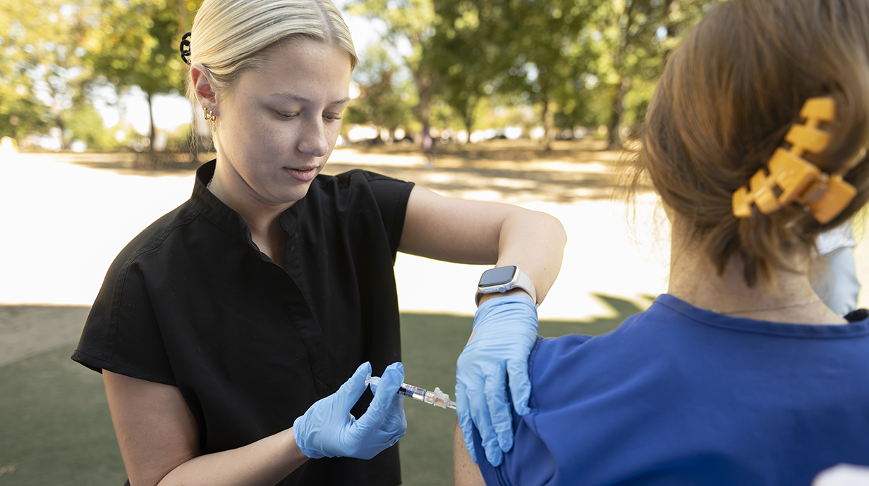 A young woman uses a syringe to give a flu shot to a patient in an outdoor setting.