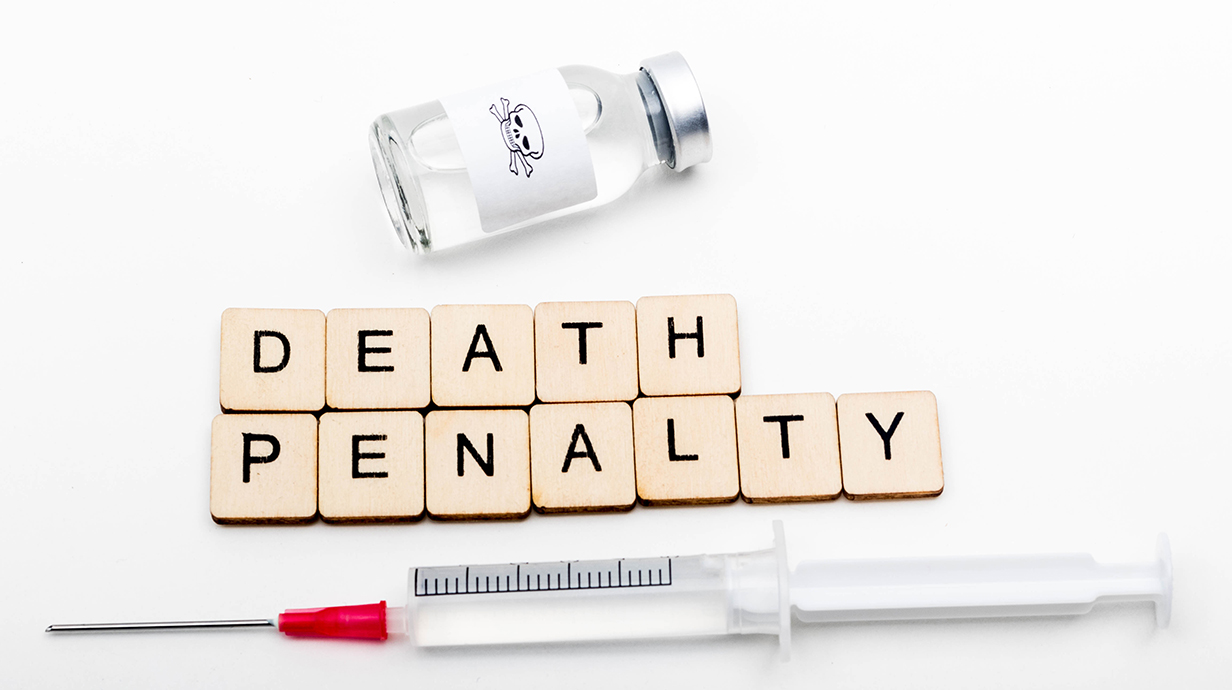 Scrabble tiles spell out 'Death Penalty' on a table with a syringe and medicine vial.