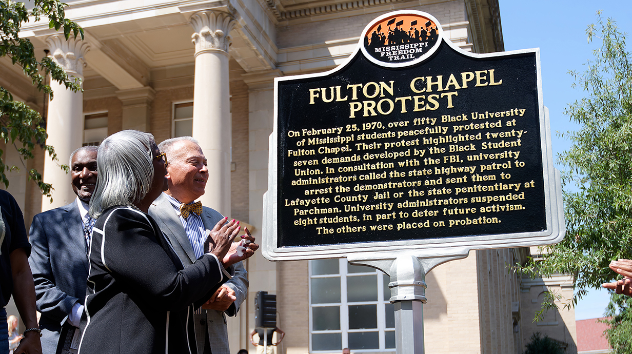 Two men and a woman applaud as a historical marker is unveiled outside a tan brick building.
