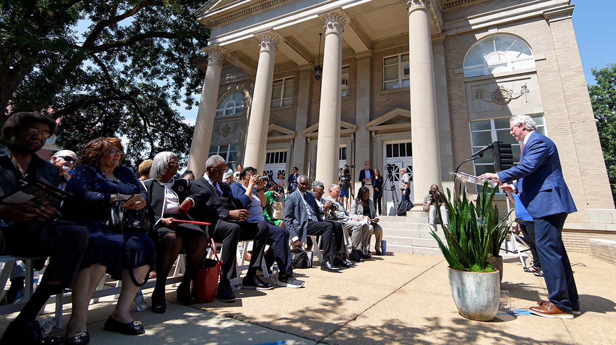 A man ata a lectern addresses a crowd seated outside a large tan brick building with white stone columns.