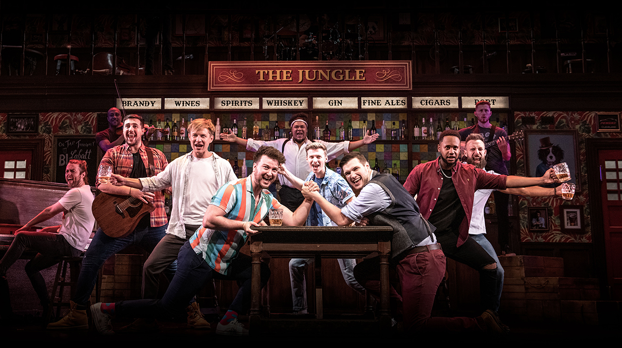 A group of men sing and dance on a stage set up as a bar.