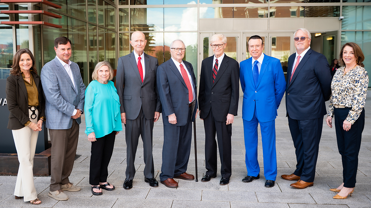 Nine people stand outside a large brick and glass building.