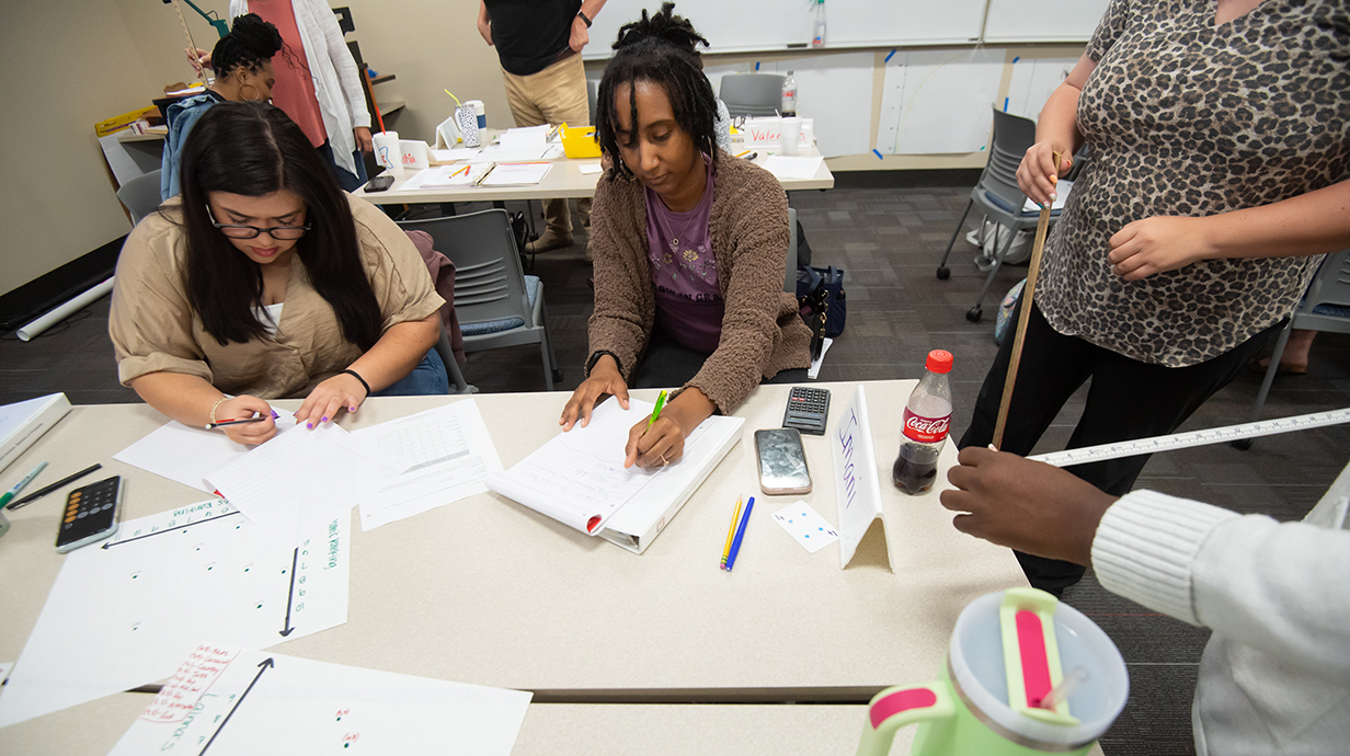 A group of women work on a class project at a table with educational materials spread out in front of them.