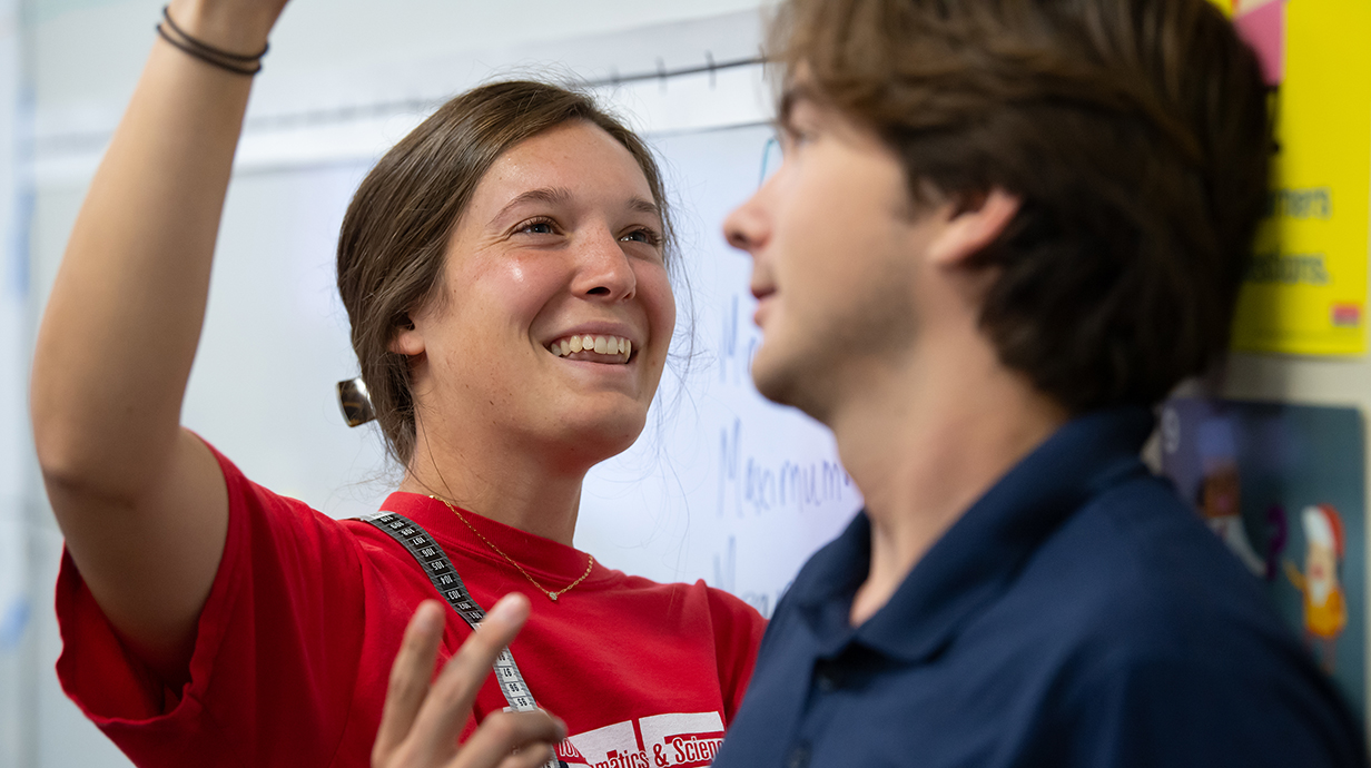 A young woman in a bright red shirt measures a young man with a ruler.