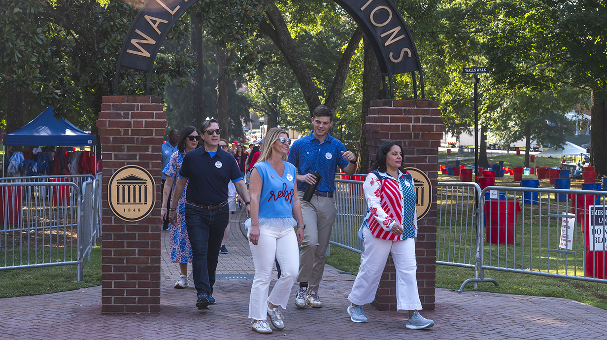 A group of people walk through an archway reading 'Walk of Champions' in a shady park.